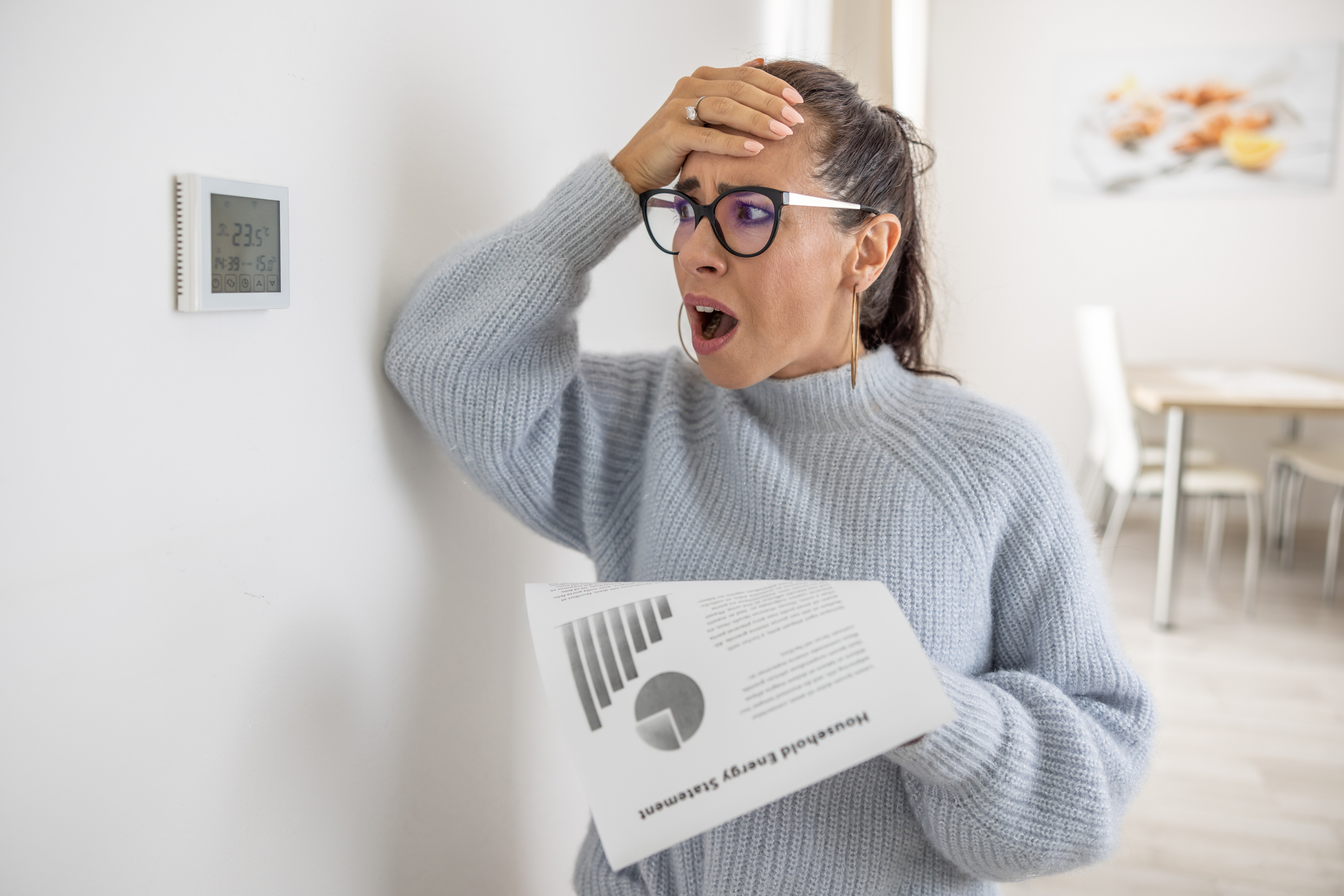 Woman cannot believe her own eyes the sum on her energy bill, looking shocked at the thermostat on the wall next to her